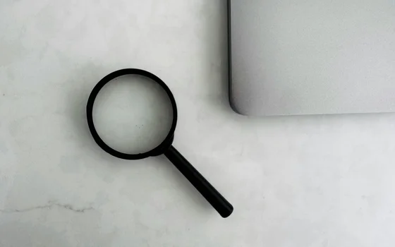 Eine Lupe liegt auf einer hellen Oberfläche neben einem Laptop. Die Lupe hat einen schwarzen Griff und eine runde Linse, die das Licht reflektiert.