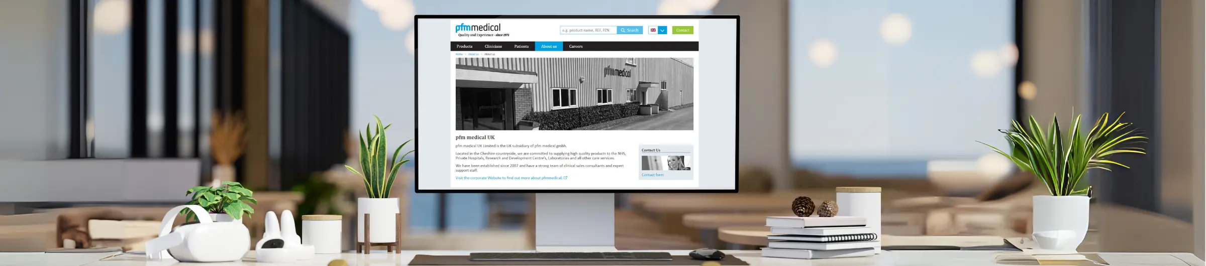 Ein Monitor zeigt die Webseite von pfm medical UK an, die Informationen über das Unternehmen und seine Produkte enthält. Im Vordergrund befinden sich einige dekorative Pflanzen und Büroartikel auf einem Tisch.