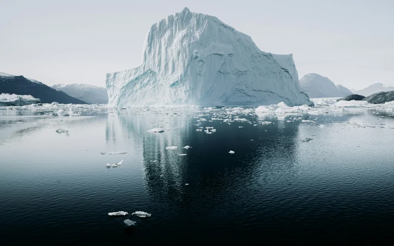 Ein riesiger Eisberg thront auf ruhigem Wasser, umgeben von treibenden Eisscherben. Im Hintergrund sind schneebedeckte Berge zu sehen. Das Wasser reflektiert die kalte Atmosphäre der arktischen Landschaft.