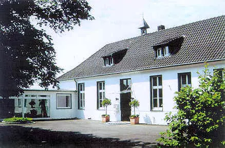 Ein zweigeschossiges, weißes Gebäude mit einem schwarzen Satteldach, zwei Fensterreihen und einem zentralen Eingang. Auf beiden Seiten des Eingangs stehen Pflanzen in Töpfen. Der Außenbereich ist gepflastert und hat einige Bäume im Hintergrund.