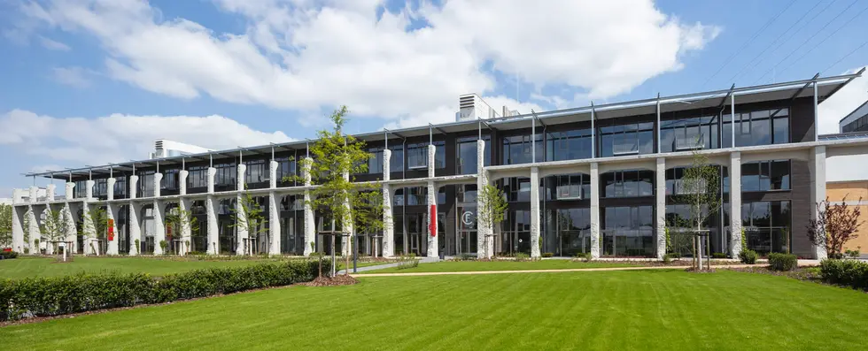 Das Bild zeigt das Arkadengebäude des Factory Campus in Düsseldorf mit großen Fenstern, umgeben von gepflegtem Grün und Büschen. Der Himmel ist größtenteils blau mit einigen Wolken.