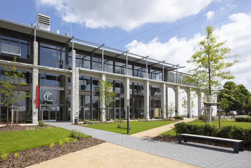 Fassade Factory Campus, Düsseldorf Ein modernes, mehrstöckiges Gebäude mit großen Fenstern und einem Eingangsbereich. Im Vordergrund befinden sich gepflegte Grünflächen mit Bäumen und Konturen. Ein grauer Gehweg führt zum Eingang, wo eine farbige Kennzeichnung sichtbar ist. Der Himmel ist freundlich mit einigen Wolken.