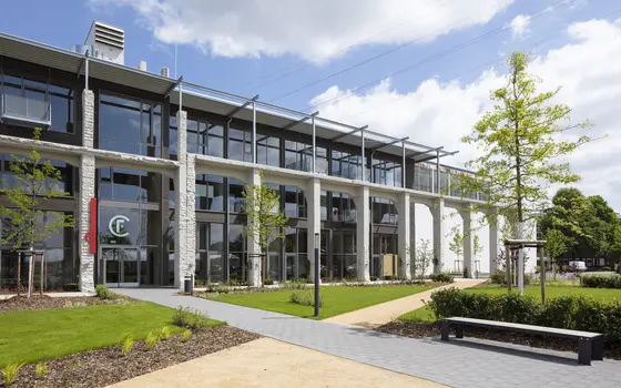 Fassade Factory Campus, Düsseldorf Ein modernes, mehrstöckiges Gebäude mit großen Fenstern und einem Eingangsbereich. Im Vordergrund befinden sich gepflegte Grünflächen mit Bäumen und Konturen. Ein grauer Gehweg führt zum Eingang, wo eine farbige Kennzeichnung sichtbar ist. Der Himmel ist freundlich mit einigen Wolken.
