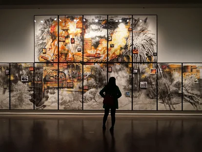Eine Betrachterin steht vor einem großen Wandgemälde, das als Collage aus vielen einzelnen Bildern in lebhaften Farben und kräftigen Pinselstrichen gestaltet ist. Das Kunstwerk zeigt dramatische Elemente wie Feuer und Rauch sowie verschiedene kleinere Bilder, die thematisch integriert sind.